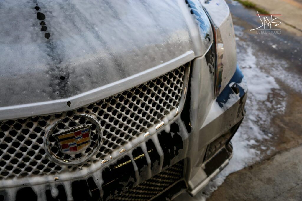 A silver Cadillac's front end covered in soapy foam, being washed outdoors.