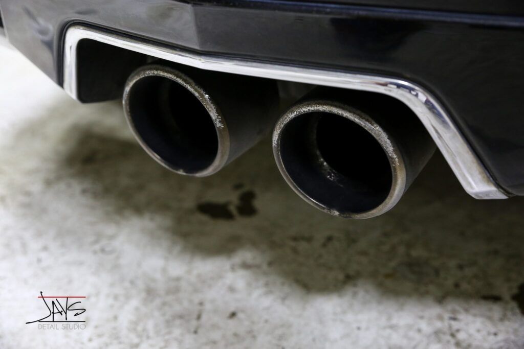 Close-up of a car's dual exhaust pipes, black and rusty, framed with a chrome border.
