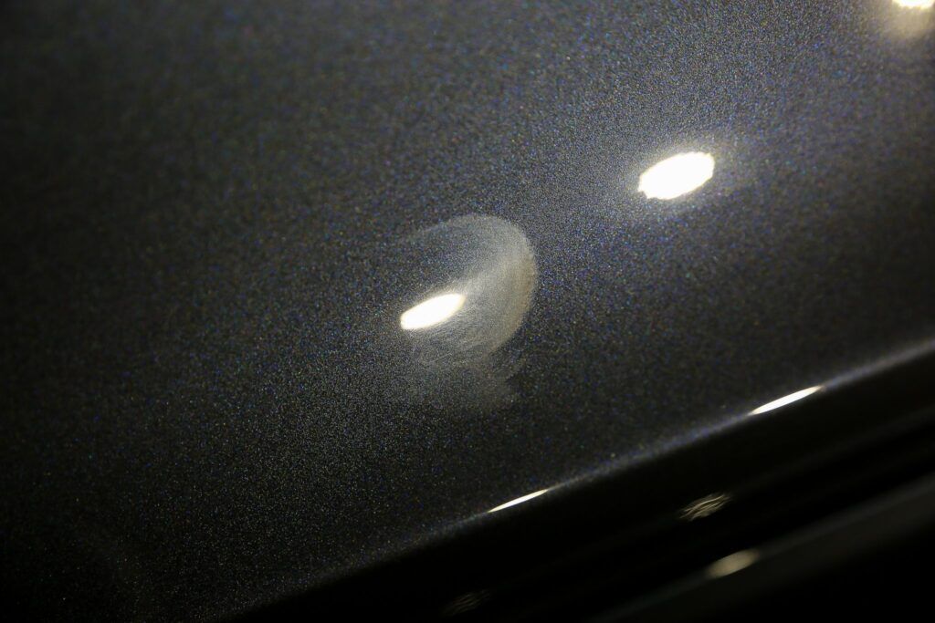 Close-up of a dark grey car surface with a water spot reflecting bright light, surrounded by fine paint particles.