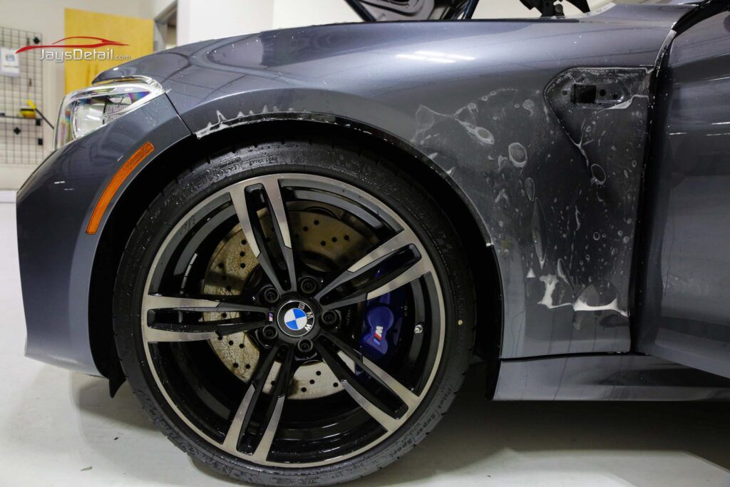 Gray BMW M car front, wheel visible; protective film partially peeled off.