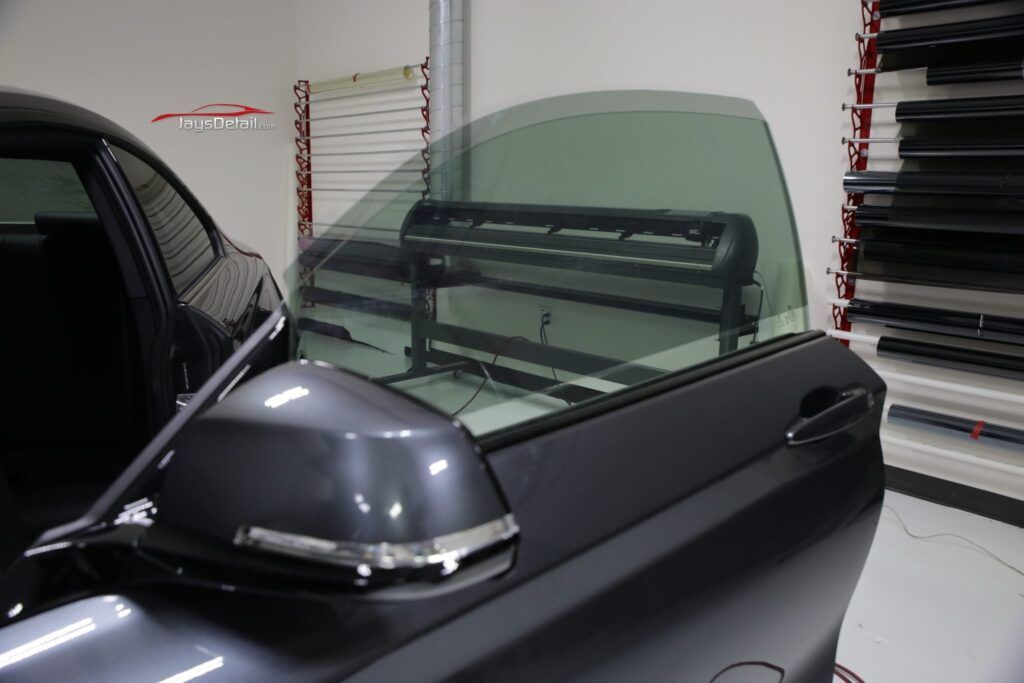 Tinted car window in a workshop, with a side view mirror and cutting machine visible.