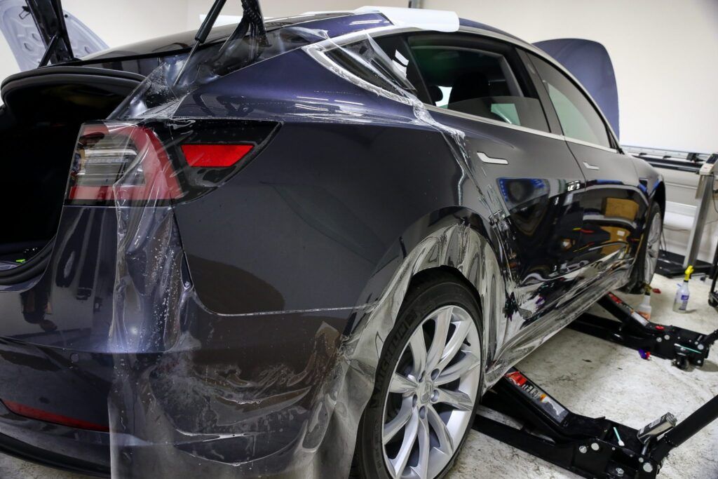 Dark blue Tesla car being wrapped in protective film in a garage.