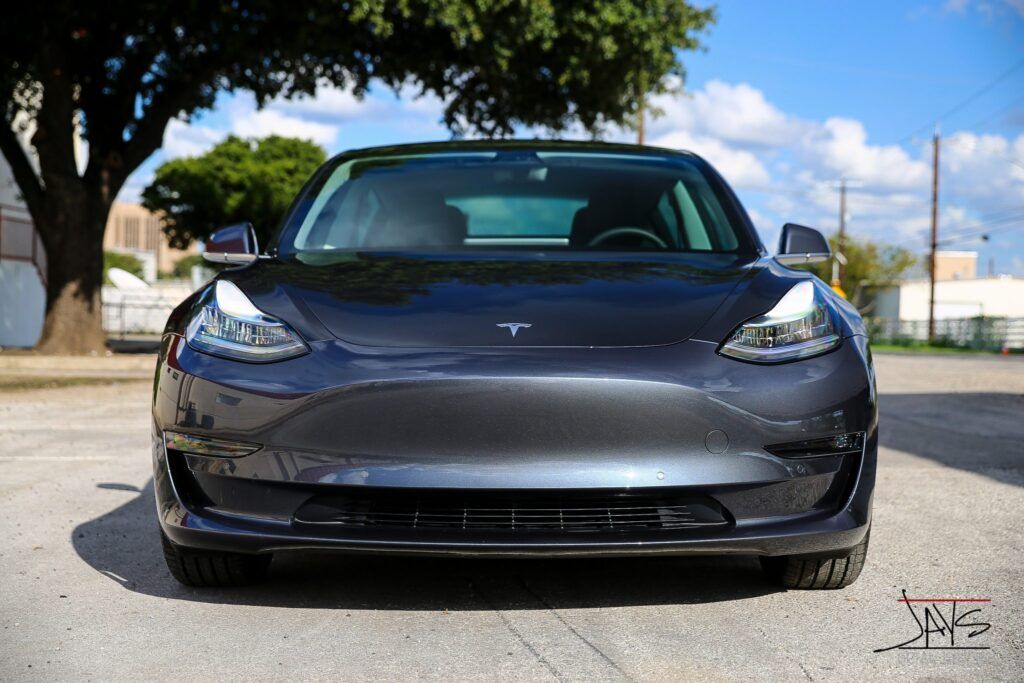 Dark gray Tesla Model 3 electric car, frontal view, parked on pavement, sunny day.