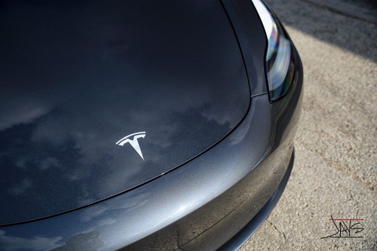 Close-up of a gray Tesla car's hood with the Tesla logo visible, reflecting the sky.
