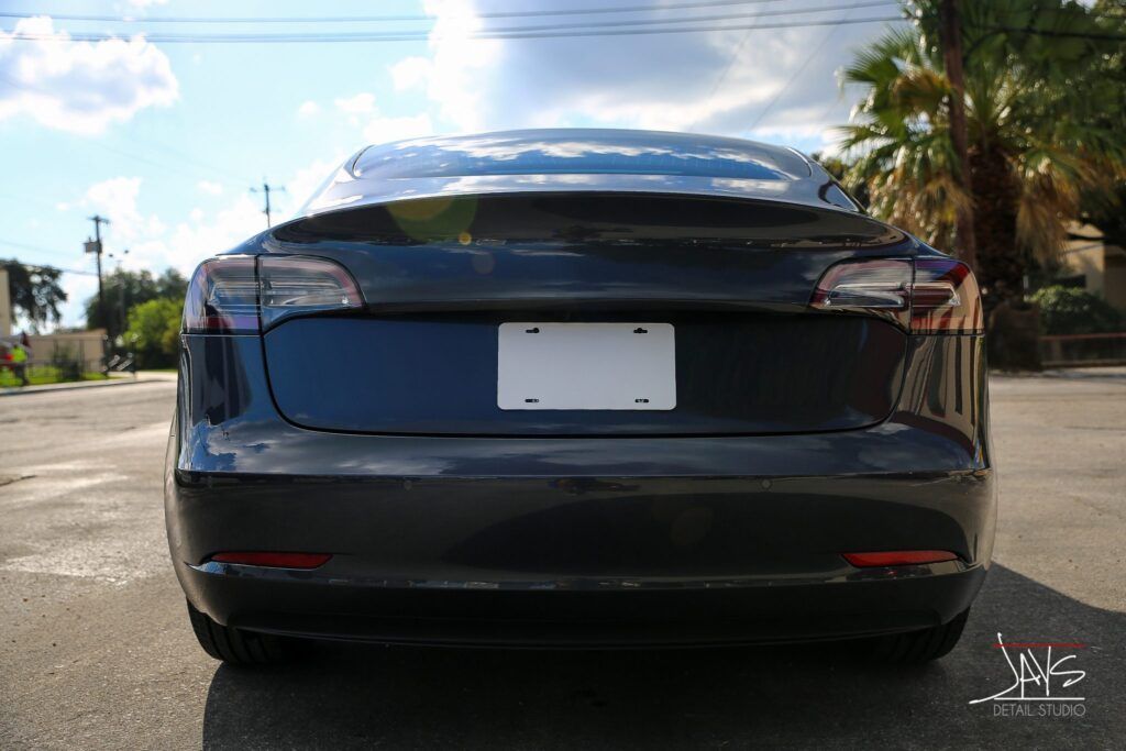 Rear view of a dark gray Tesla sedan on a street, license plate obscured, red taillights.