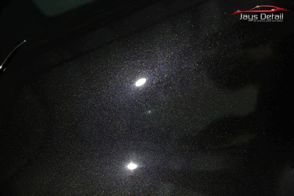 Close-up of a black car hood with numerous visible imperfections highlighted by bright overhead lights.