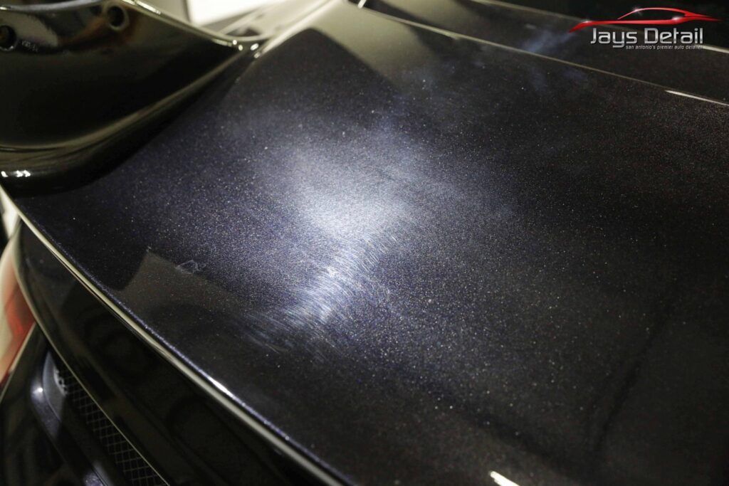 Close-up of a dark car's hood, showing swirls and scratches under a bright light.