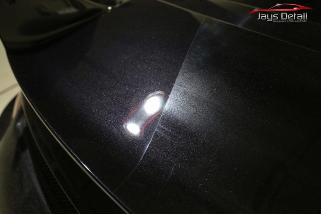 Close-up of dark car paint showing swirl marks illuminated by a bright light; Jay's Detail logo visible.