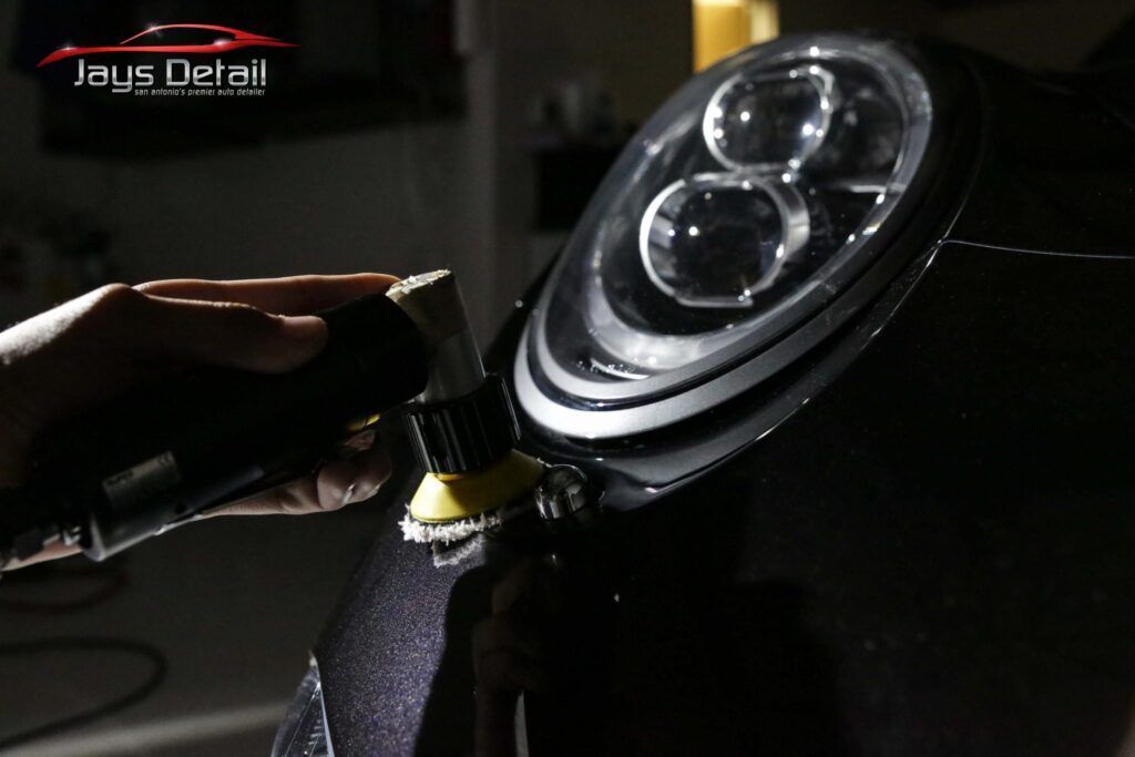 Detailer polishes a car headlight on a black vehicle with a power tool and yellow pad.