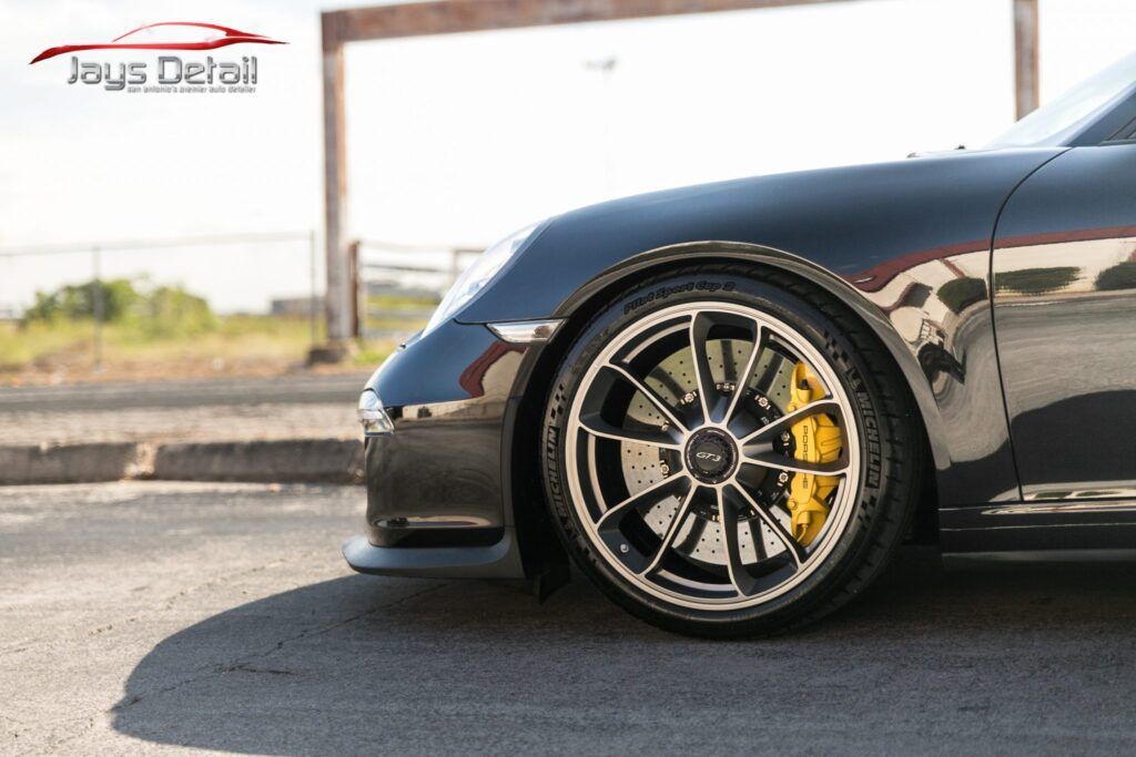 Close-up of a black Porsche's front wheel with yellow brake calipers, parked on the street.