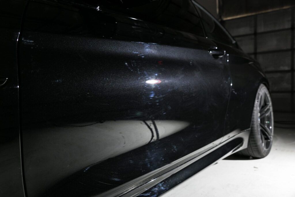 Black car door and side panel; showing scuffs and scratches, under a bright light.
