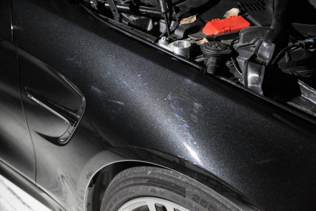 Black car fender with open hood, engine visible. Tire and rim in view.