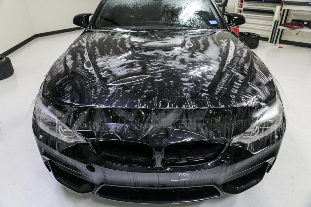 Black BMW covered in clear paint protection film in a shop.