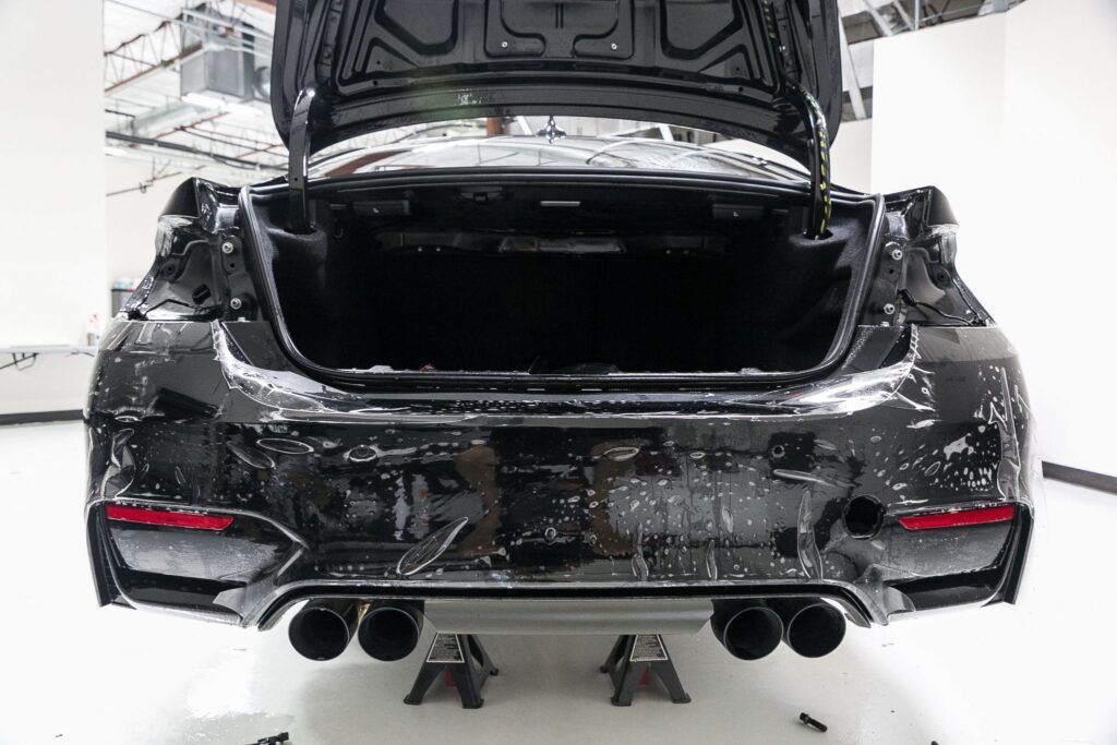 Black BMW car, rear view, trunk open, covered in foam, on jack stands.