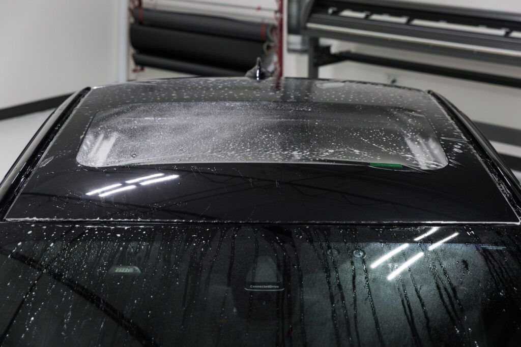 Black car roof with water droplets, window tinting in progress, inside a workshop.