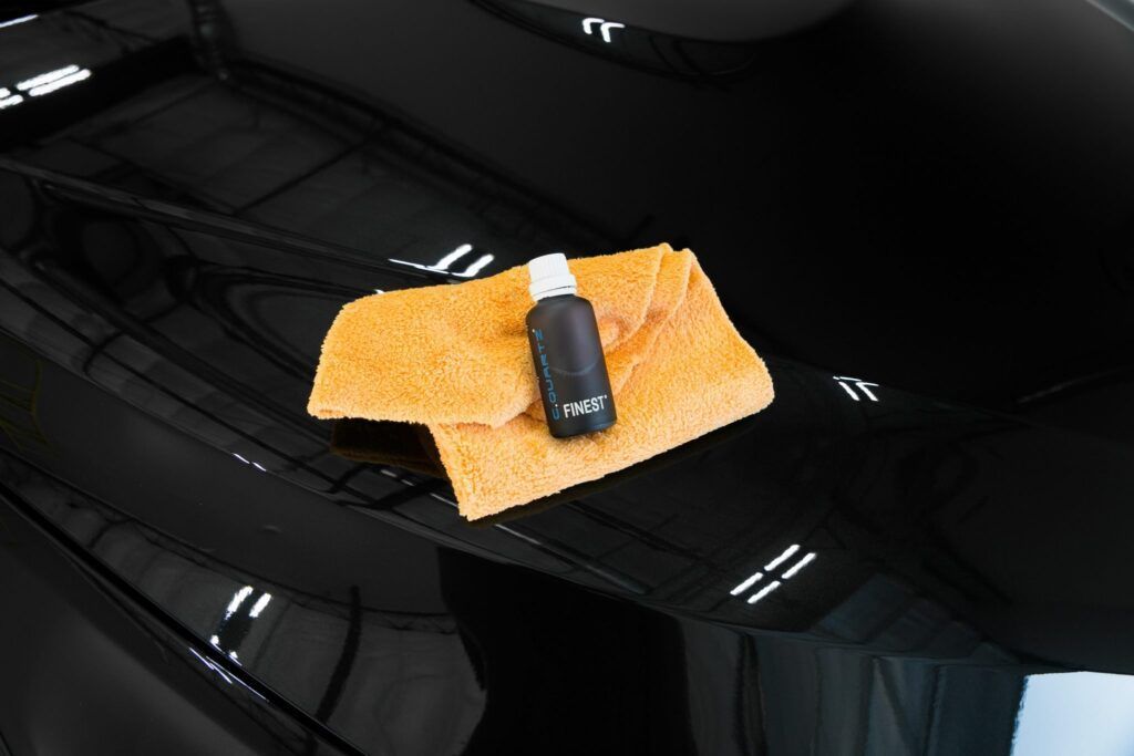 Black car hood with bottle and orange microfiber cloth for detailing.