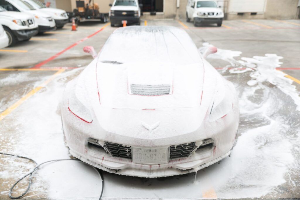A white sports car covered in white foam, being washed in a parking lot.