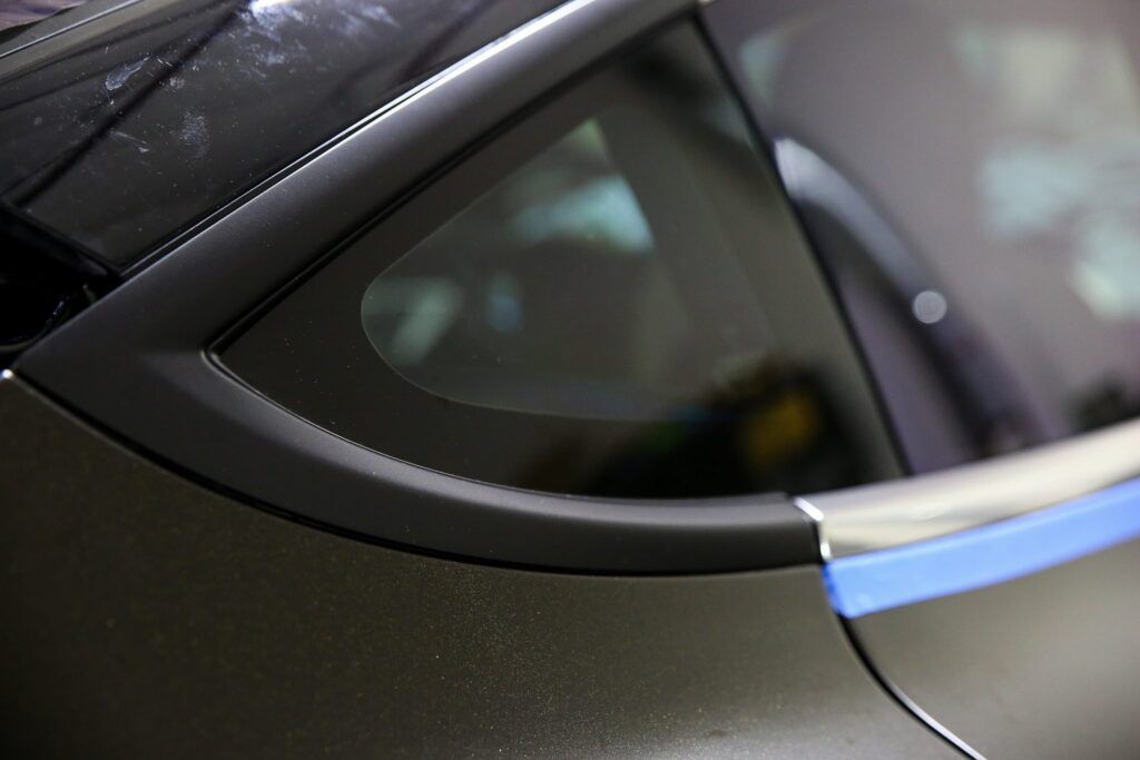 Close-up of a car's black window and frame with blue tape, showing detailing work.