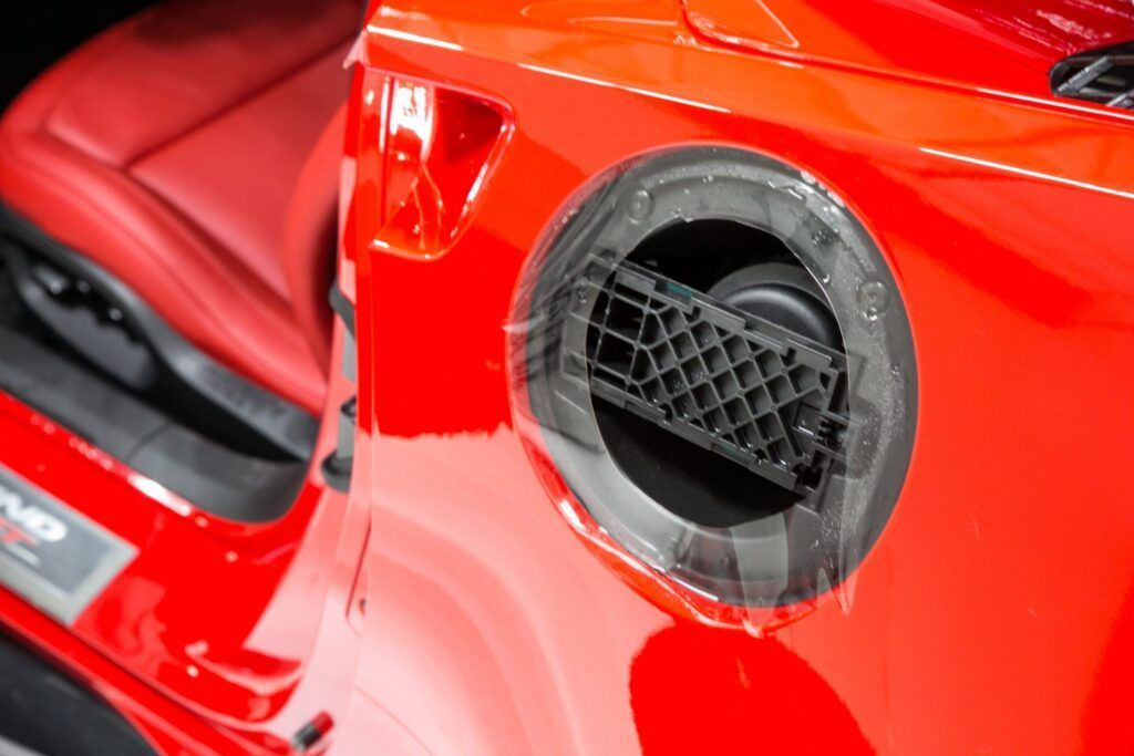 Red car's fuel door, with a dark plastic grate visible within the opening, contrasting with the glossy paint.