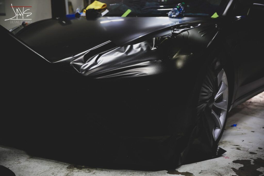 Black car being wrapped with black film in a garage, the front end is visible.