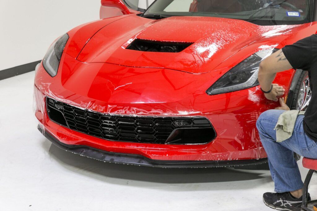 Person applying clear protective film to the red front bumper of a sports car in a well-lit garage.