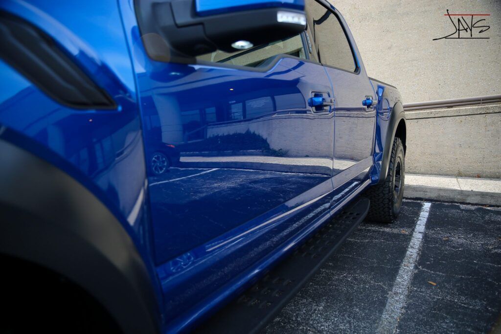 Blue Ford truck parked next to a building, angled view, showing side panel, mirror, and running board.