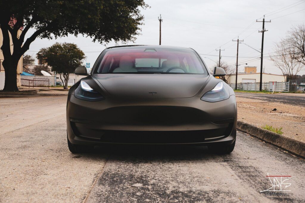 Dark matte Tesla car parked on a paved road; overcast day.