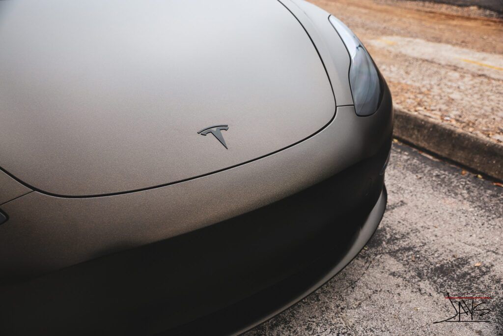 Close-up of a dark gray Tesla car's hood with the Tesla logo.