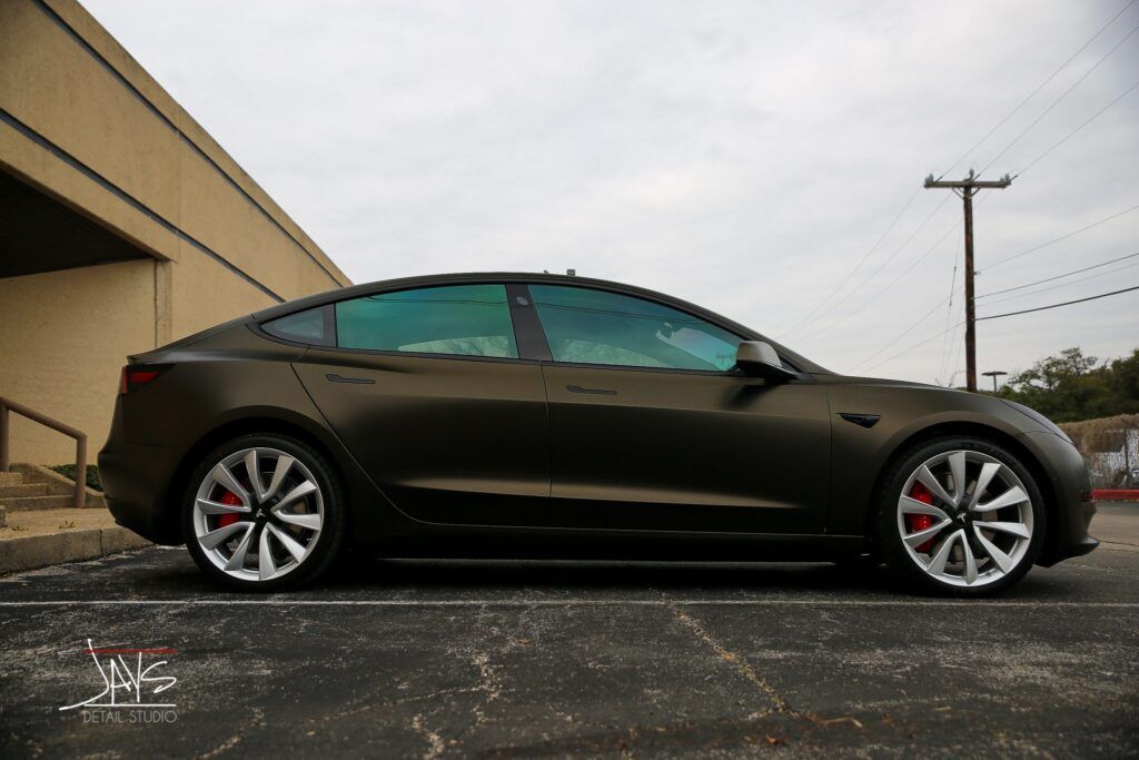 Dark brown Tesla Model 3 with tinted windows and red brake calipers, parked next to a building and road.