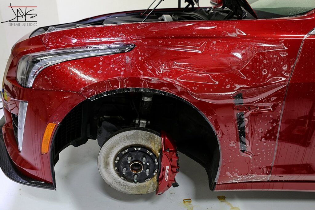 Red car's front fender with protective film being applied; garage setting.