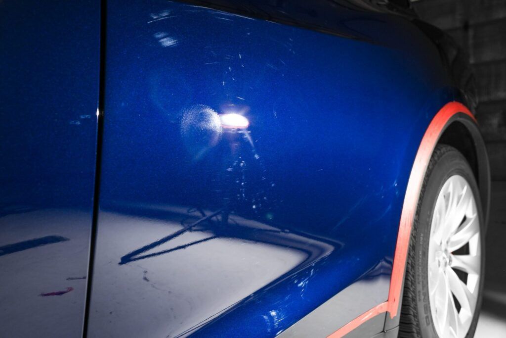 Blue car door with swirls and scratches reflecting light, detailing in progress.