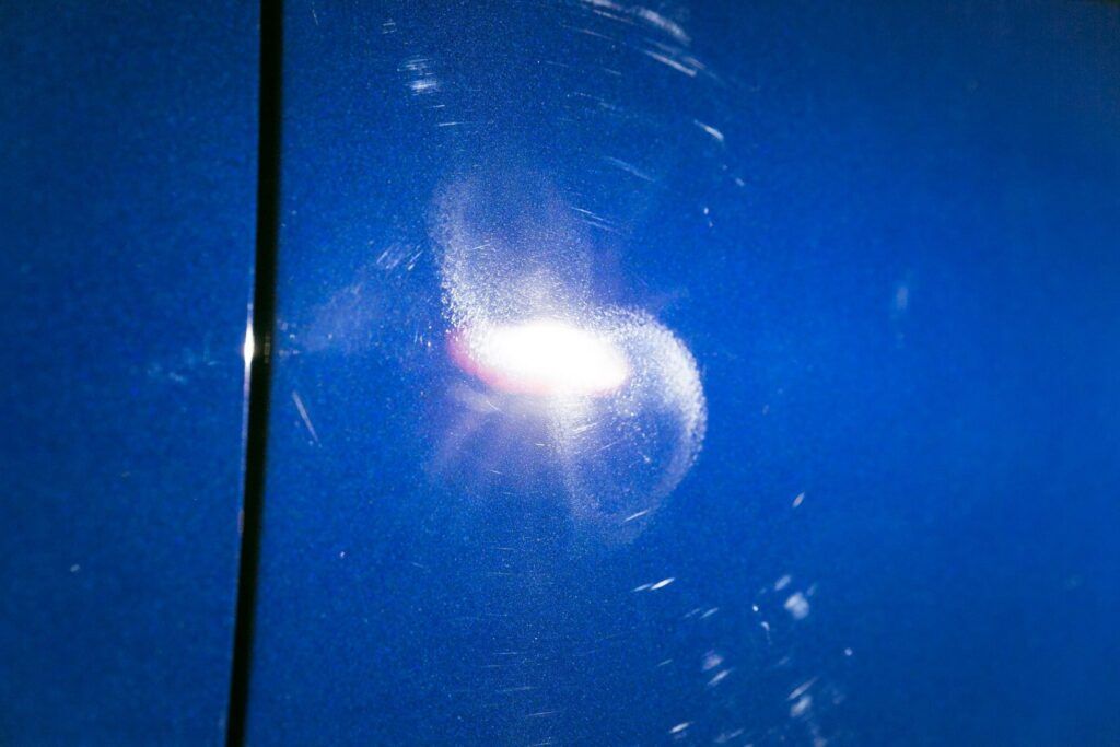 Blue car paint with circular scratches and a bright light reflection.
