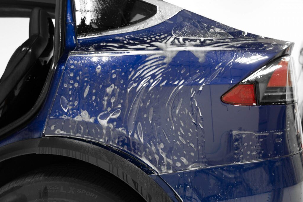 Blue car's rear quarter panel being covered in protective film, with visible soap and water.