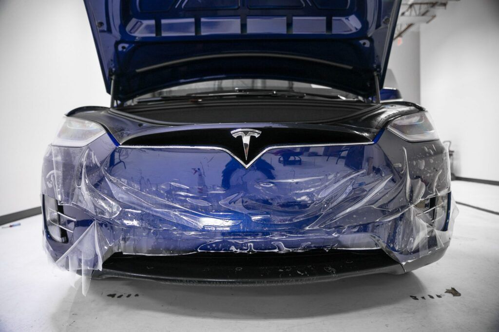 Blue Tesla car with hood open, front covered in protective wrap, indoors.