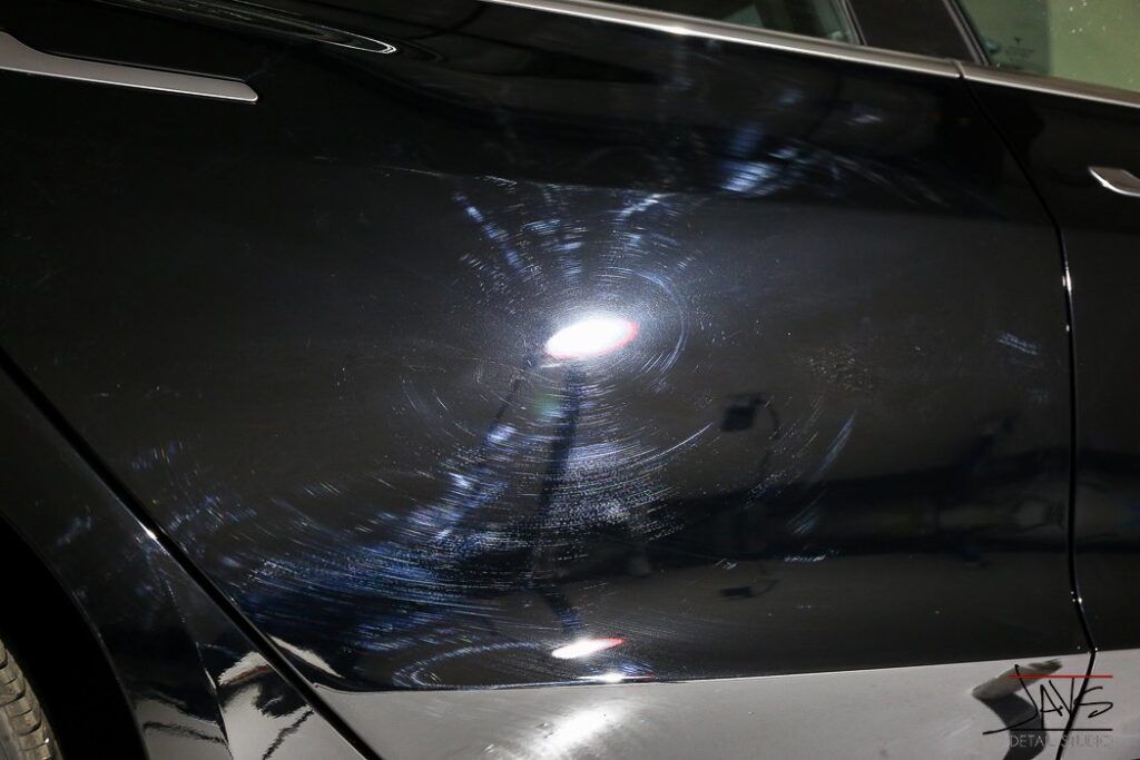 Black car door with swirling scratches and light reflections.