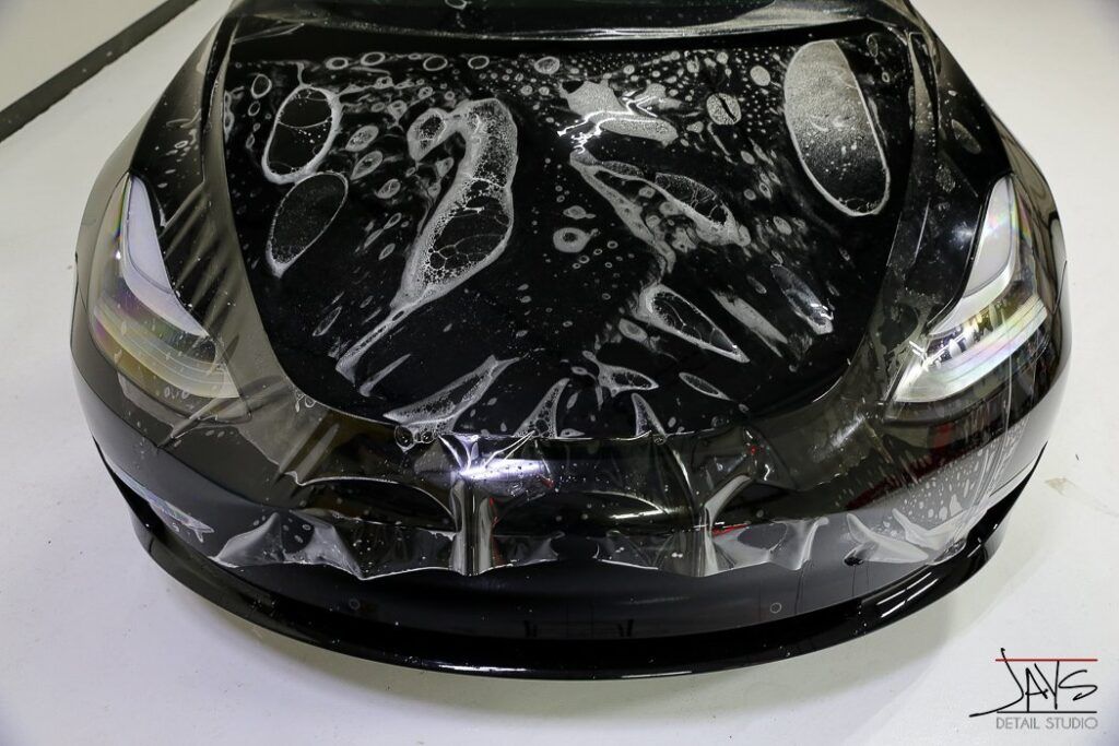 Car hood with protective film being applied, reflecting overhead lights.