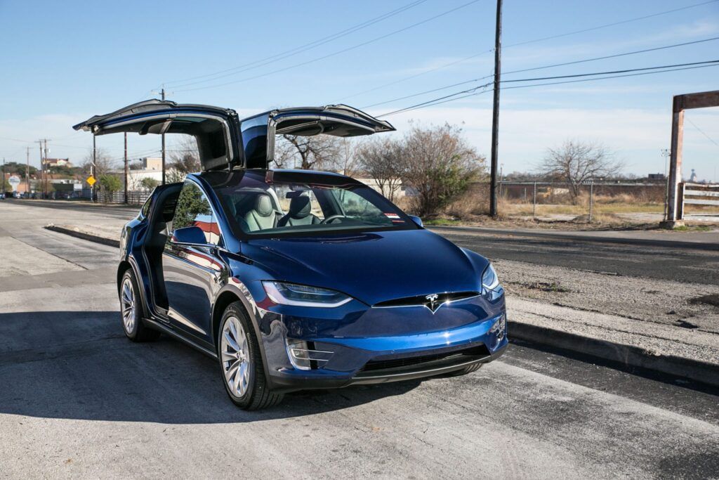 Blue Tesla Model X with open falcon-wing doors on a gravel road.