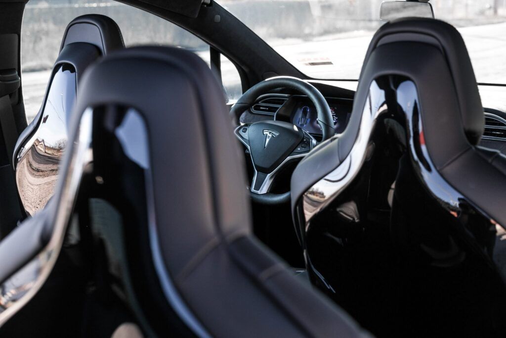 Interior view of a Tesla car showing black leather seats with chrome accents and the steering wheel.
