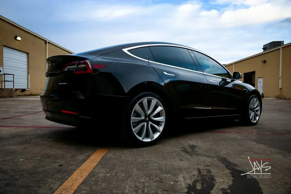 Black Tesla car parked on a paved lot. The car is positioned between two tan buildings under a cloudy sky.