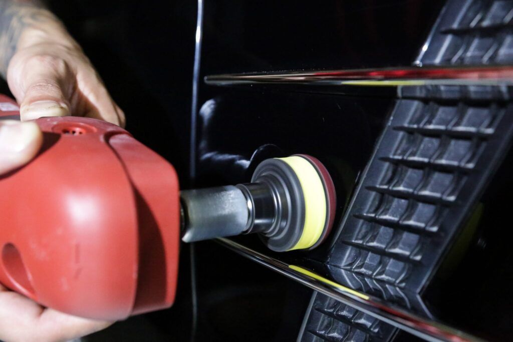 A person uses a red power polisher on a black car grill to remove imperfections.