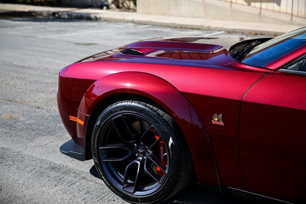 Red Dodge Challenger's front quarter panel with black wheels and a hood scoop, parked on asphalt.