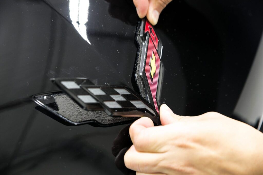 Hands peeling off a red and black emblem from a black car. The emblem has a checkered flag design.