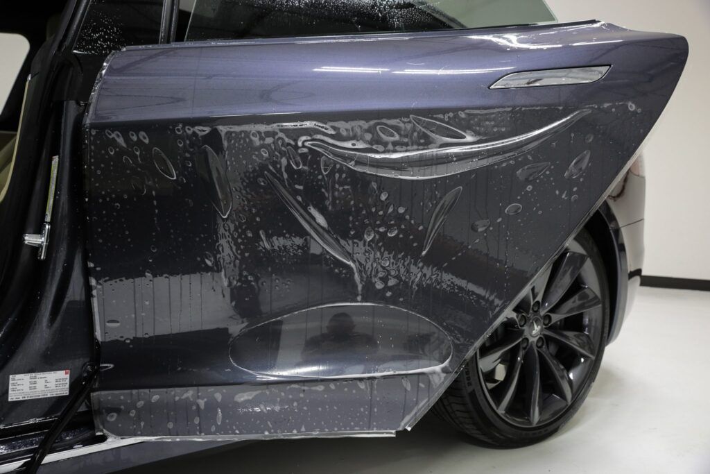 Car door with clear film being applied, dark blue paint, in a workshop setting.