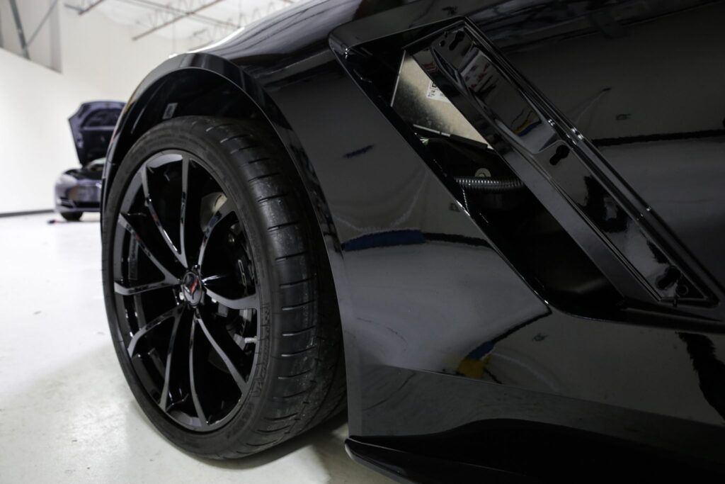 Black sports car's front quarter panel, wheel, and air vent, parked indoors on a white floor.