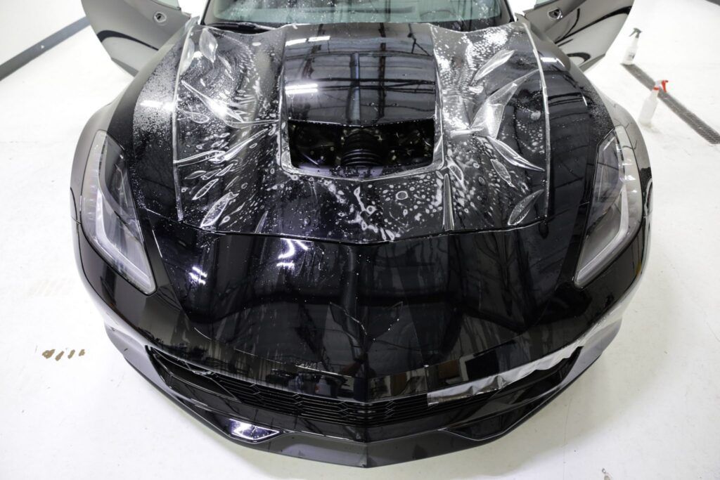 Black Corvette with protective film applied to the hood. Doors open, indoors, white floor.