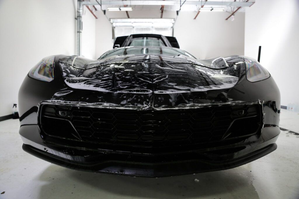 Black Corvette sports car covered in foam, being washed in a bright garage.