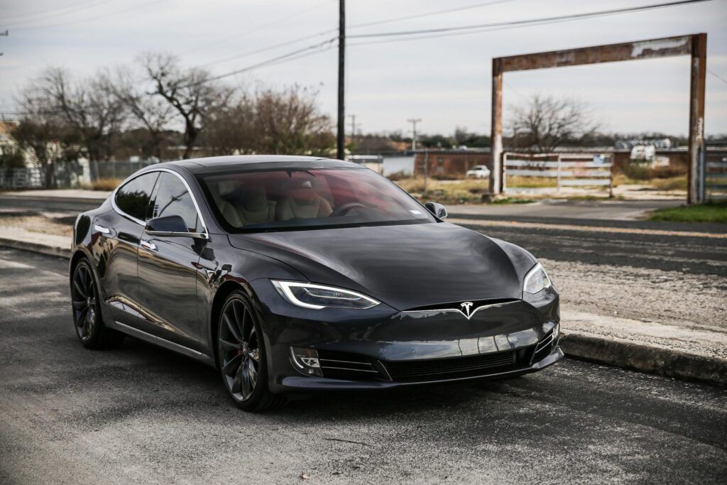 Dark gray Tesla Model S parked on a road, with a rusty metal structure in the background.