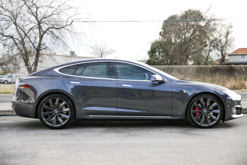 Gray Tesla Model S parked on a street with bare trees and a low fence in the background.