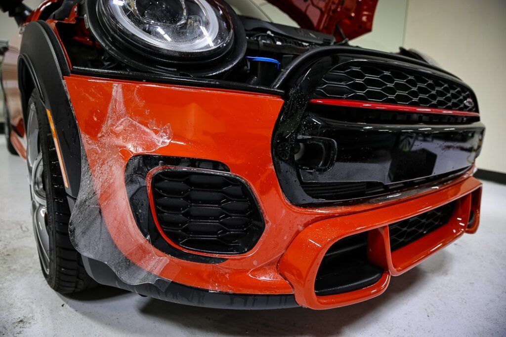 Close-up of a red and black Mini Cooper's front bumper, with exposed engine and open hood.