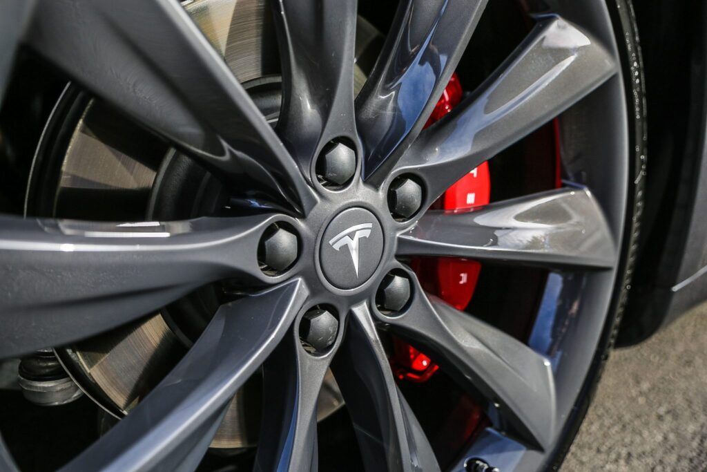Close-up of a Tesla car wheel. Gray alloy with black lug nuts and a red brake caliper.
